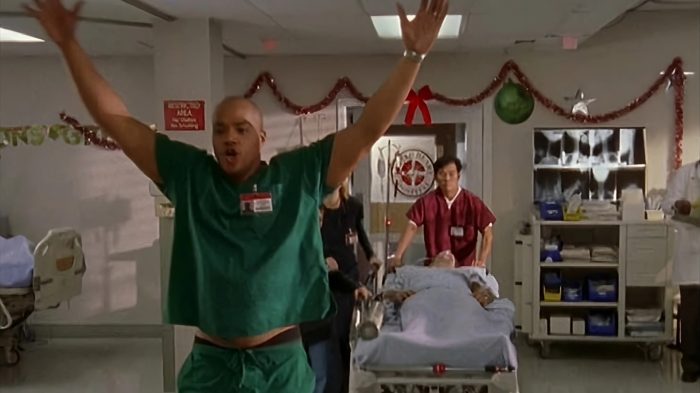 Turk standing in the Sacred Heart hospital ward with his arms raised in a celebratory pose next to Christmas decorations.